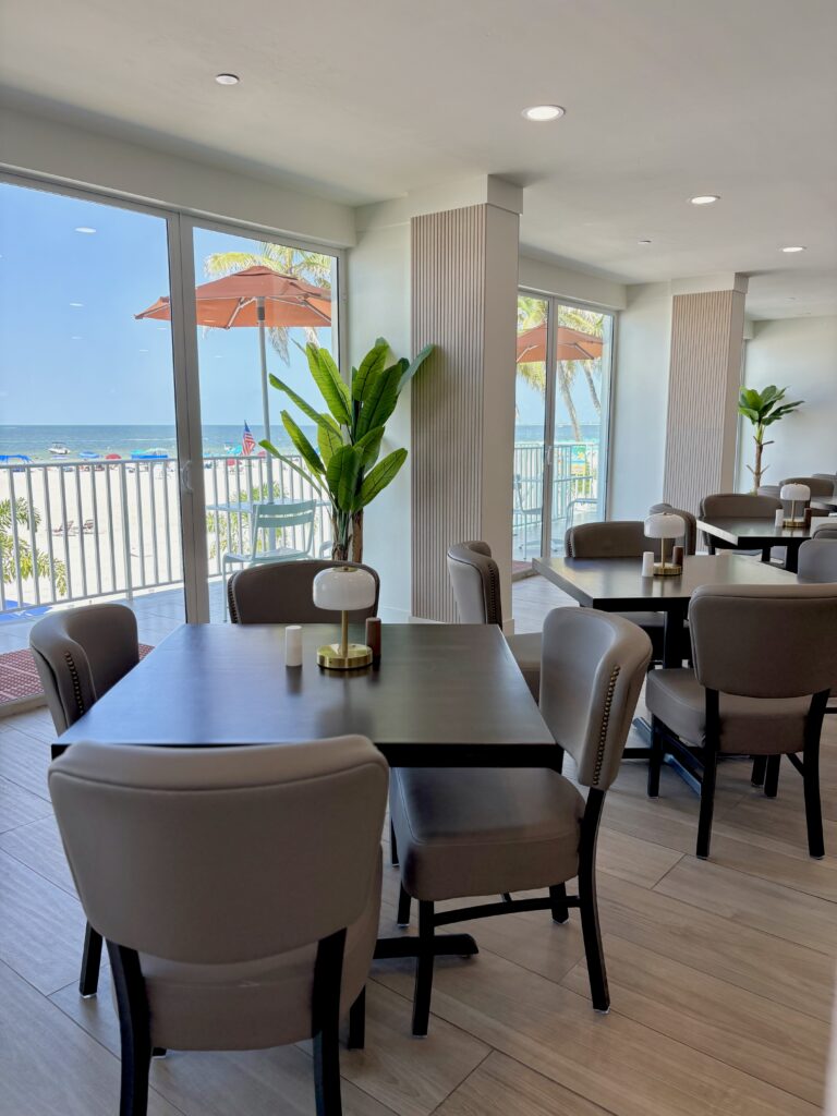 Casual indoor restaurant that overlooks the beach, with tables and chairs and beach-inspired design.