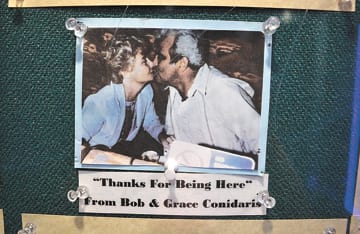 Polaroid picture of man and woman sitting at table, kissing. Picture is hung on a board and has a quote of 'Thanks for being here from Bob and Grace Conidaris" underneath the picture
